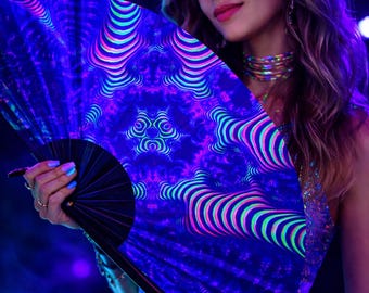 May include: A vibrant, patterned hand fan with a black handle. The fan features a repeating design of curved lines in neon blue, pink, and green against a dark blue background. The fan is held open, showcasing its intricate design.
