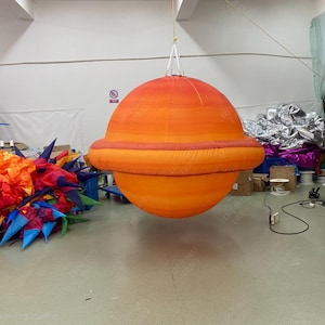 Op de afbeelding: Een oranje opblaasbare planeet met een ring, hangend aan het plafond. De planeet heeft een kleurenschema met een verloop, van een lichter oranje aan de bovenkant naar een donkerder oranje aan de onderkant. De ring is een donkerdere tint oranje.