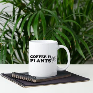 Peut inclure: Une tasse à café en céramique blanche avec du texte noir qui dit "COFFEE & PLANTS" et un petit dessin au trait noir d'une plante. La tasse est posée sur un carnet noir.