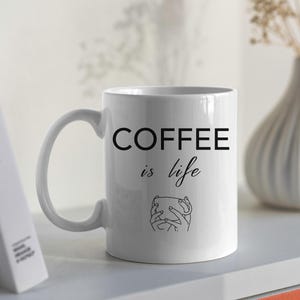 Pode incluir: Caneca de café de cerâmica branca com texto preto que diz "COFFEE is life" e um desenho de linha de uma mão segurando uma xícara de café.