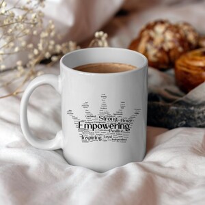 Puede incluir: Taza de cerámica blanca con un diseño de corona negra hecho de palabras como "fuerte", "valiente", "empoderamiento", "inspirador" e "independiente". La taza está llena de un líquido marrón y está sobre una superficie blanca.