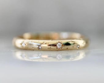 Op de afbeelding: Een gouden ring met een hemels ontwerp. De ring heeft kleine, sprankelende diamanten en stervormige gravures. De band is glad en gepolijst en reflecteert licht. De ring is een sieraad.