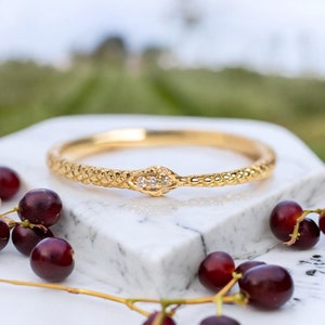 May include: A gold bangle bracelet with a textured snake design. The snake's head has two small, clear gemstones. The bracelet is on a white marble surface, with dark red berries in the foreground.