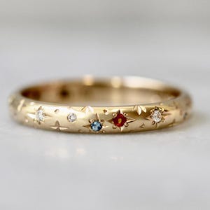 May include: A gold ring with a celestial design. The ring features small star and leaf patterns, and is adorned with several gemstones, including clear, blue, and red stones. The ring is a delicate piece of jewelry.