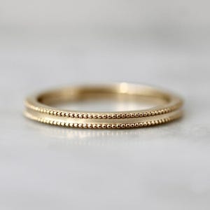 Puede incluir: Un anillo de oro con un diseño de cuentas. El anillo tiene una banda lisa y pulida con una fila de pequeñas cuentas a lo largo de los bordes superior e inferior. El anillo es probablemente de oro y tiene un acabado brillante.