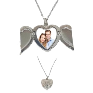 May include: A silver heart-shaped locket necklace with a photo of a couple inside. The locket opens to reveal the photo and has wing-like details on either side. The necklace is displayed on a white background.