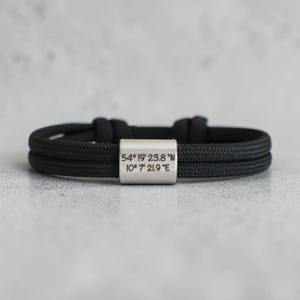 Könnte beinhalten: Ein schwarzes Seilarmband mit einem silberfarbenen rechteckigen Verschluss. Auf dem Verschluss sind die Koordinaten "54° 19' 23.8" N 10° 7' 21.9" E" eingraviert. Das Armband liegt auf einer hellgrauen Oberfläche.