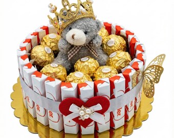 May include: A decorative gift arrangement resembling a cake, featuring a gray teddy bear wearing a gold crown, surrounded by chocolate candies. The arrangement includes a red heart with a white bow and pearl accents, and a gold butterfly.