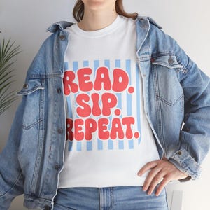May include: A white t-shirt featuring a light blue vertical striped square background with the red text READ. SIP. REPEAT. in a bold, rounded font. The t-shirt is worn under a light blue denim jacket with metal buttons and blue denim jeans.
