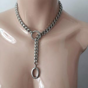 Puede incluir: Un collar de cadena plateada con un colgante ovalado grande. El collar está hecho de eslabones de cadena gruesos e interconectados. El colgante cuelga de una cadena más pequeña que se conecta a un anillo más grande. El collar se exhibe en un maniquí.