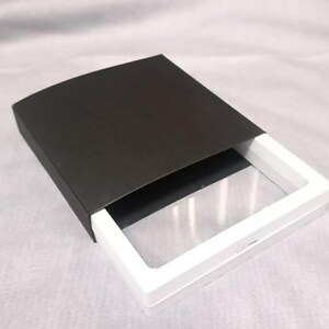 以下が含まれることがあります： 白いフレームと透明な窓、そして黒いスライド式の外カバーを備えた長方形のディスプレイケース。黒いカバーから白い内側のトレイを部分的に引き出すことができる構造で、透明な表示エリアが露出するようになっています。