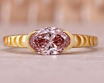 May include: A gold ring featuring a large, oval-cut pink gemstone. The band has a unique design with cut-out crescent shapes on each side of the gemstone. The ring is set against a soft, neutral background.