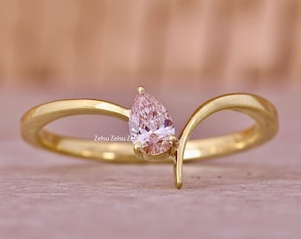 May include: A gold ring with a pear-cut, pale pink gemstone. The ring has a distinctive, curved design, with the gemstone set at the top. The ring is set against a soft, blurred background.