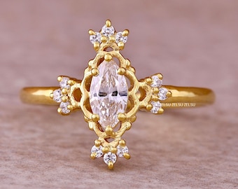 May include: A gold-colored ring with a marquise-cut diamond center stone. The ring features a decorative filigree design with small, round, clear gemstones and a crown-like detail at the top. The ring is set against a neutral background.