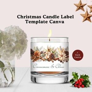 May include: A white candle with a cinnamon and clove scent, decorated with a festive winter-themed label. The label features pine cones, cinnamon sticks, and other wintery elements. The candle is in a clear glass jar and is lit.