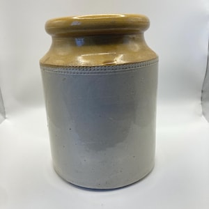 May include: A stoneware jar with a light grey body and a tan-coloured rim and top. The jar has a cylindrical shape with a slightly wider base and a decorative band of small circles around the middle. The jar is likely vintage.