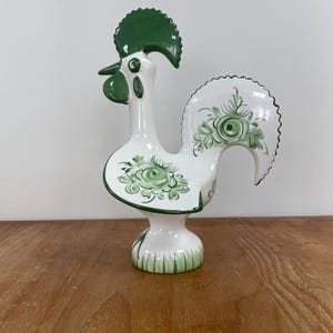 Puede incluir: Una figura de gallo de cerámica blanca con cresta y barbillas de color verde sólido. El cuerpo y la cola están decorados con motivos florales pintados a mano en verde. La base de la figura está acentuada con un diseño de hierba pintado en verde.