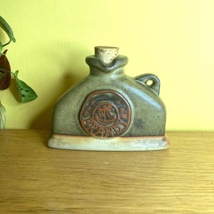 May include: A ceramic flask with a cork stopper. The flask is olive green with a circular, reddish-brown emblem. It has a small handle and sits on a rectangular base. The flask is displayed on a wooden surface.