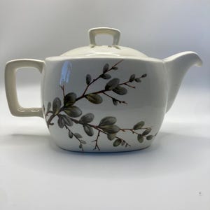May include: Cream-colored ceramic teapot with a matching lid. The teapot features a curved handle and spout, and is decorated with a delicate floral branch design in shades of green and brown. The teapot is designed for brewing tea.