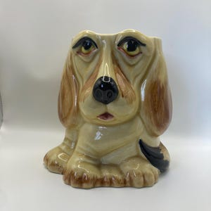 Op de afbeelding: Een keramische hondenpot met een crèmekleurige basis en bruine oren. De hond heeft een zwarte neus, zwarte ogen en een kleine rode mond. De pot is ontworpen om planten of bloemen in te zetten.