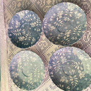 Könnte beinhalten: Vier blaue und weiße Untertassen mit floralem Muster. Die Untertassen haben ein detailliertes Blumendesign mit Blättern und Blumen. Der Hintergrund ist hellblau. Die Untertassen sind auf einer weißen Spitzentischdecke angeordnet.