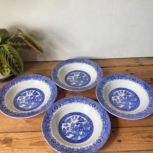 May include: Four white and blue Willow pattern soup bowls. Each bowl features a central blue landscape scene surrounded by a blue patterned border. The bowls are arranged on a wooden surface.