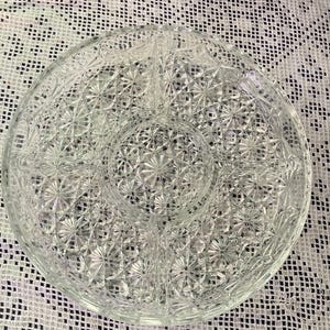 Könnte beinhalten: Eine klare, runde Servierschale mit Sternmuster und Unterteilungen. Die Schale besteht aus Glas und hat vier Fächer, ideal zum Servieren von Snacks oder Vorspeisen. Die Schale hat einen Durchmesser von etwa 30 cm.