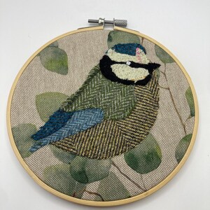 以下が含まれることがあります： 木製のフレームに手作りの鳥の刺繍。鳥は緑、青、茶色のテクスチャード加工された生地を使用し、白黒の顔があります。背景はベージュ色の生地に緑の葉の模様が施されています。