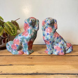 Pode incluir: Duas estatuetas de cão em cerâmica com padrões florais em rosa, azul e verde. Os cães estão sentados, com as orelhas caídas. As estatuetas estão sobre uma superfície de madeira.