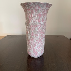 May include: A cylindrical vase featuring a scalloped rim and a textured surface with a mottled pattern of soft pink, white, and light grey colours. The body of the vase has vertical ridges that create a subtle, undulating effect across the exterior.