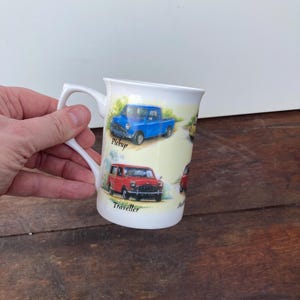 May include: A white ceramic mug decorated with illustrations of classic cars, including a blue pickup and a red Traveller. The mug has a curved handle and is set against a light background.