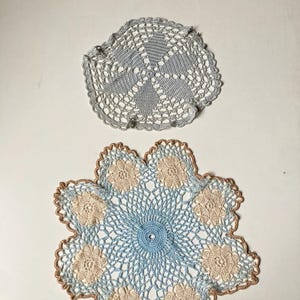 Può includere: Due centrini all'uncinetto fatti a mano. Il centrino superiore è grigio con un bordo smerlato. Il centrino inferiore è blu con accenti floreali beige e un bordo smerlato marrone. Questi pezzi decorativi sono perfetti per aggiungere un tocco di fascino vintage.