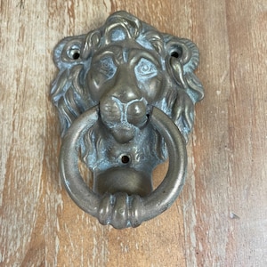 May include: A brass-toned metal door knocker shaped like a lion head with a circular ring held in its mouth. The lion features a detailed mane and facial features, with three visible screw holes for mounting on a door surface.