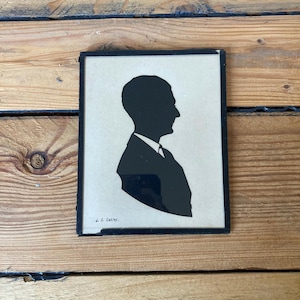 May include: A framed black silhouette portrait of a man in profile, wearing a suit and tie. The silhouette is set against a cream-coloured background. The frame is black, and the artist's signature, "J. S. Oakley," is visible.