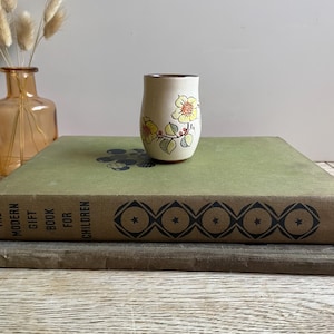 Può includere: Un piccolo bicchiere in ceramica bianco sporco con un motivo floreale giallo e rosso. I fiori sono accompagnati da foglie e steli verdi. Il bordo del bicchiere ha una fascia marrone. Il bicchiere è su un libro verde.