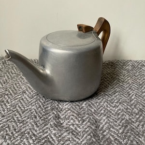 May include: A vintage silver-colored teapot with a wooden handle and lid knob. The teapot has a curved spout and a rounded body. The handle and lid knob are made of light-colored wood. The teapot is sitting on a gray patterned surface.