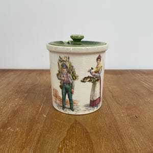 May include: A cylindrical ceramic jar with a crackled cream glaze and a green lid. The exterior features a colourful illustration of two figures in historical attire, one carrying a large bundle of sticks and the other holding a small hammer.
