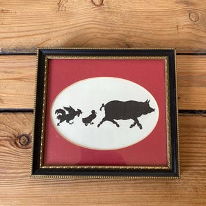 Può includere: Opera d'arte incorniciata con sagome nere di un gallo, un'anatra e un maiale che corrono su un ovale bianco contro uno sfondo rosso. La cornice è nera con un bordo interno dorato, su una superficie di legno.