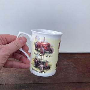 Op de afbeelding: Witte keramische mok met illustraties van vintage tractoren. De mok heeft een gebogen handvat en toont de tekst "David Brown Vak IC" en "Fordson Model 'N'". De tractoren zijn afgebeeld in rood en groen op een gele achtergrond.