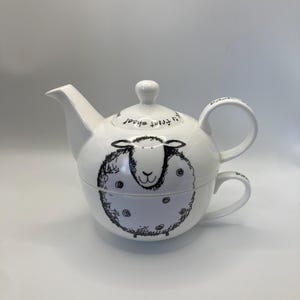 Op de afbeelding: Witte keramische theepot en theekopjes set met een schapenontwerp. De theepot heeft een tuit en een handvat, en de theekop past eronder. Het schapenontwerp is zwart en wit, met de woorden "it front sheep" op het deksel.