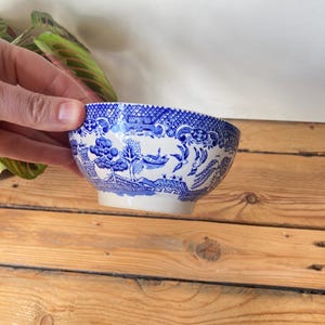 May include: A small, blue and white ceramic bowl with a detailed, traditional pattern. The bowl features a scalloped rim and a classic landscape design. The bowl is held against a wooden background.