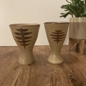 May include: Two beige ceramic cups with brown geometric designs. The cups have a wide base and a narrow top.