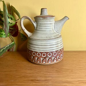 May include: A ceramic teapot with a light beige body and a brown patterned base. The teapot has a curved handle, a spout, and a lid with a small knob. The design includes horizontal lines and geometric shapes.