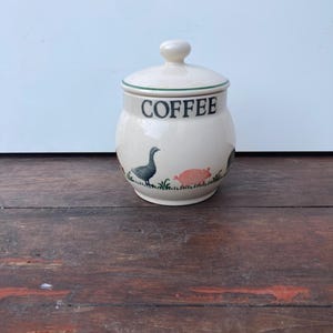 May include: Cream-colored ceramic coffee canister with a lid. The word "COFFEE" is printed in black. The canister features illustrations of a goose, a pig, and grass. The lid has a small knob.