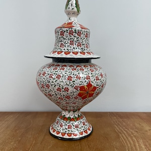 May include: A ceramic lidded jar featuring a white base with intricate hand-painted floral patterns in red, green, and black. The vessel has a bulbous body, a pedestal base, and a pointed, removable lid, all decorated with repeating botanical motifs.