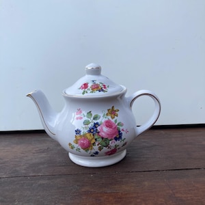 Può includere: Una teiera in ceramica bianca con coperchio, beccuccio e manico. La teiera è decorata con un motivo floreale con rose rosa, bacche blu e fiori gialli. I bordi dorati accentuano i bordi della teiera e del coperchio. La teiera poggia su una piccola base rotonda.