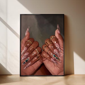 Op de afbeelding: Ingelijste kunstafdruk met een close-up van handen met lange, amandelvormige nagels. De nagels zijn nude geschilderd met zwarte en gouden accenten. Gouden ringen sieren de vingers, en de achtergrond is donker en abstract.