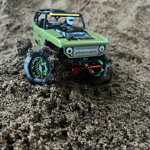 May include: A green toy truck with black wheels and a green and pink rim, driving through sand.