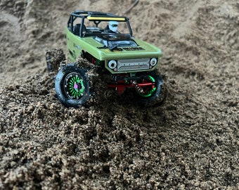 May include: A green toy truck with black wheels and a green and pink rim, driving through a pile of sand.