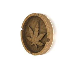 May include: A wooden ashtray featuring a carved marijuana leaf design. The ashtray is oval shaped and has a brown wood finish.